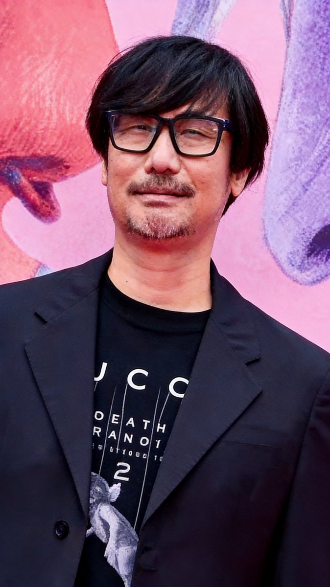 Hideo Kojima in a dark blazer in front of a pink and purple illustrated backdrop