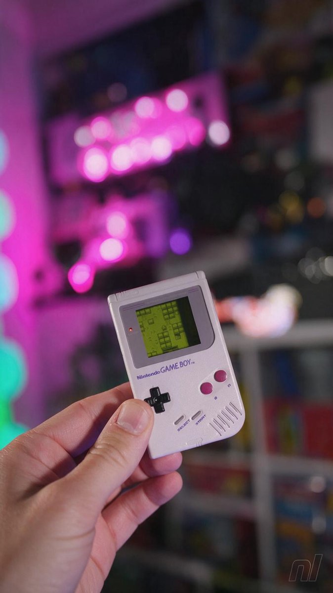 A hand holds the chunky Game Boy-shaped jukebox, its green pixel screen glowing — the whole 1996 soundtrack in one $69.99 shell