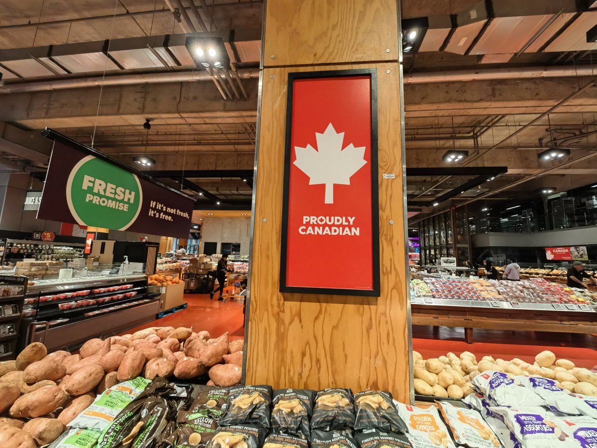 A large red Proudly Canadian sign with a white maple leaf mounted on a pillar in the produce section, beside bins of potatoes
